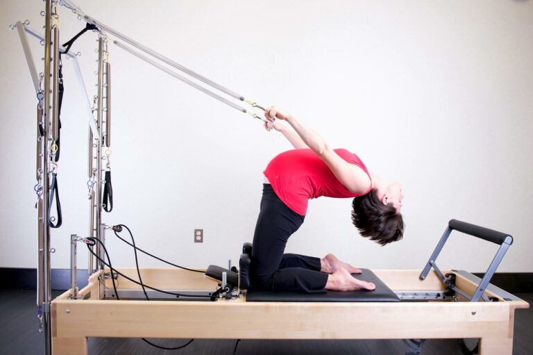 Woman performing stretching exercise on a Pilates reformer in a brightly lit gym environment.